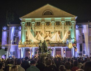 Das Deutsche Nationaltheater Weimar erstrahlt in bunten Lichtern, während eine Menschenmenge eine Veranstaltung im Freien genießt.
