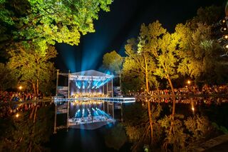 Eine lebhafte Konzertbühne erstrahlt nachts im Weimarhallenpark, umgeben von leuchtenden Bäumen und einem ruhigen Teich, der die Lichter reflektiert.